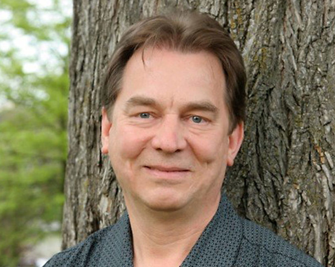 a smiling man standing in front of a pine tree