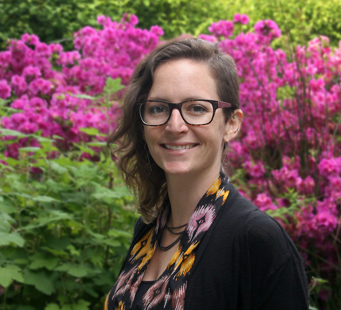 a smiling woman standing in front of a large bush with bright pink flowers,