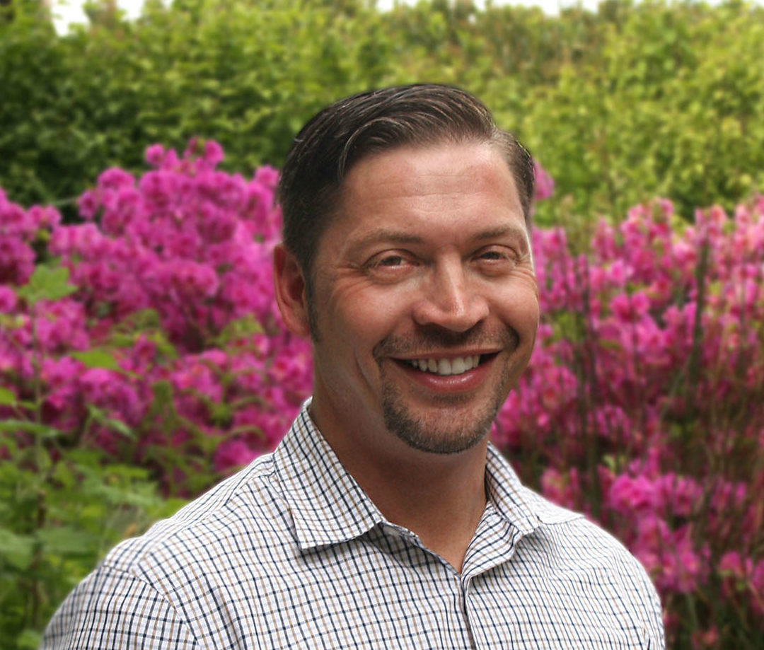 a smiling man standing in front of a large bush with bright pink flowers,