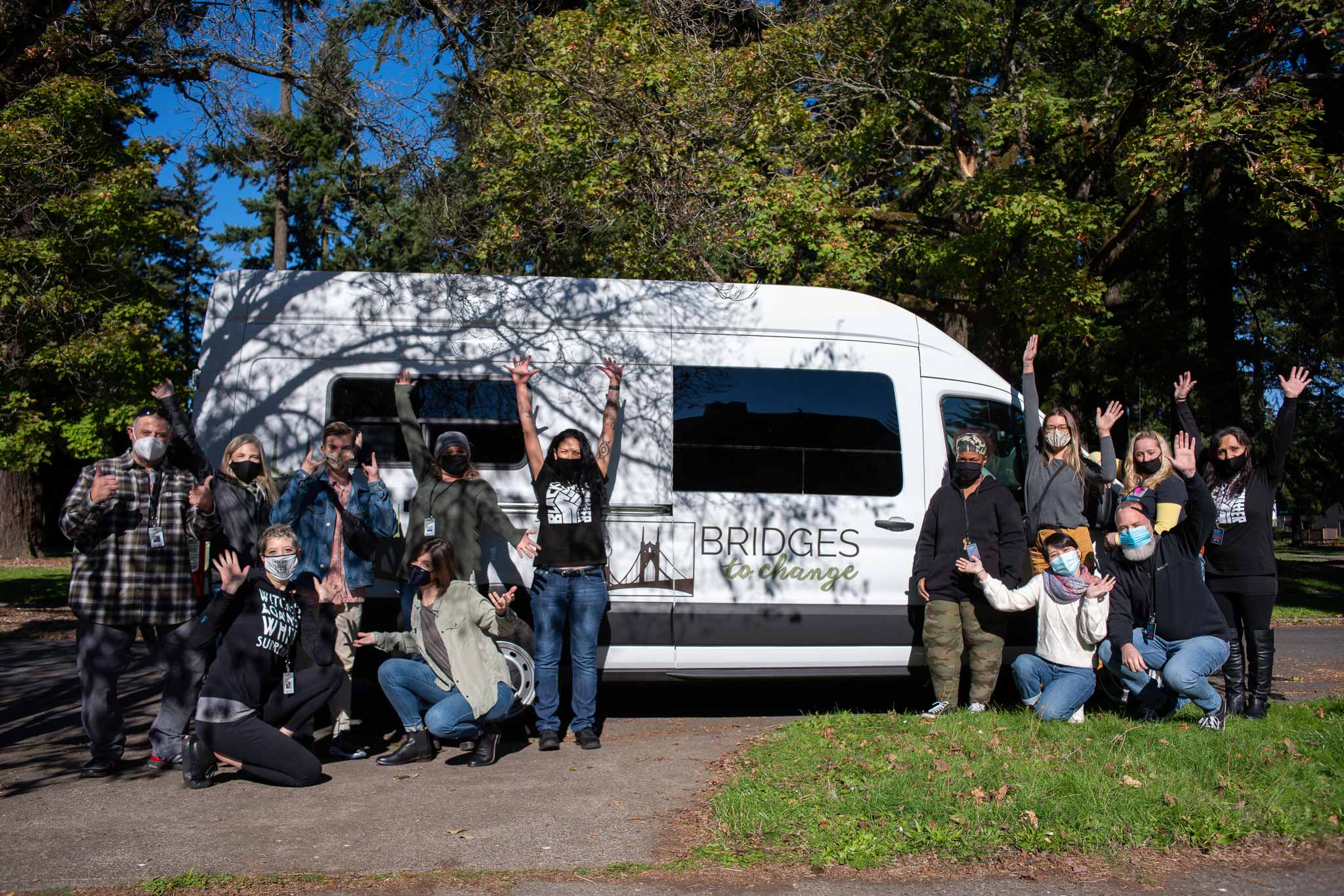 a group of people kneeling and standing while throwing their arms up in elation in front of the bridges to change community outreach sprinter van
