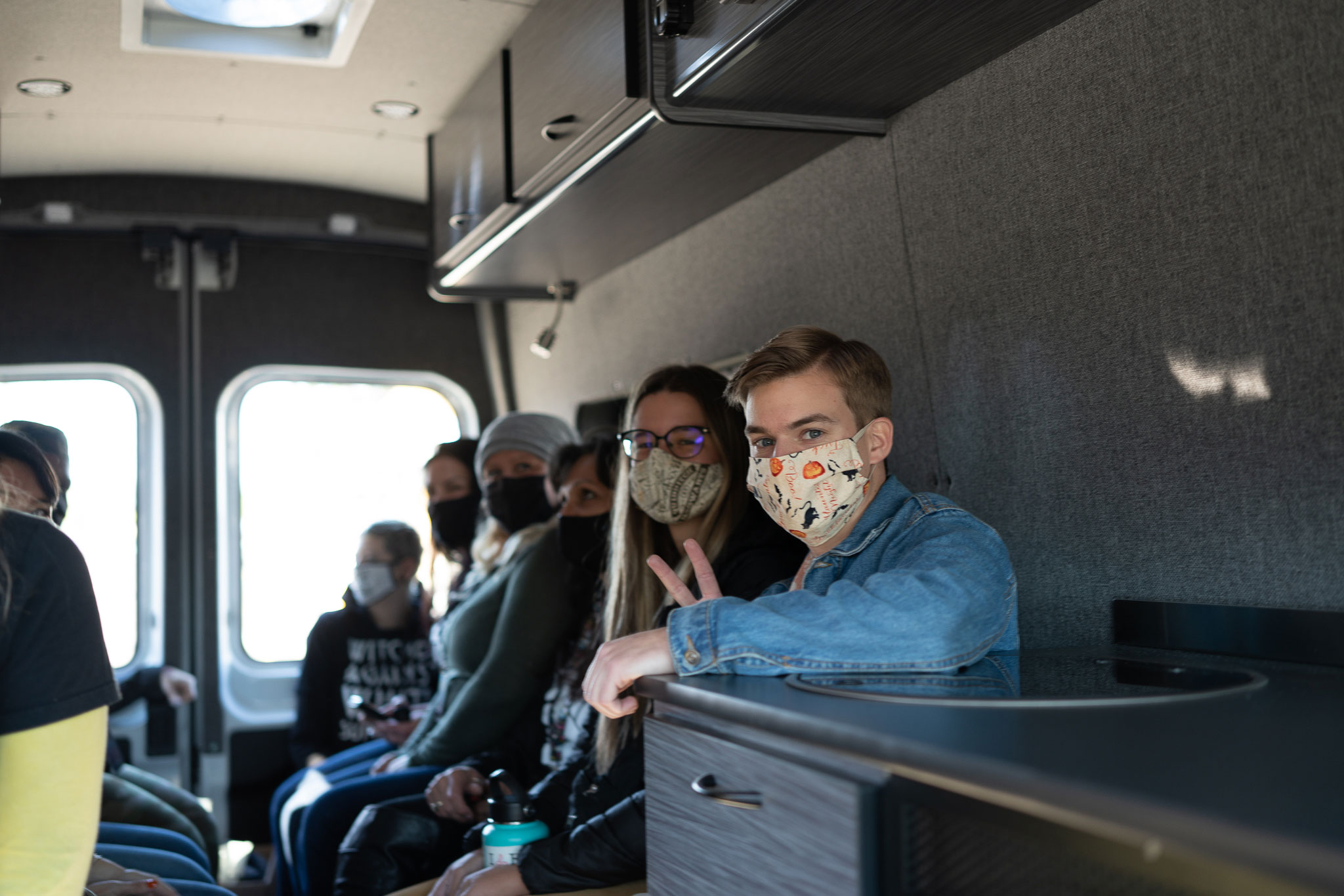 a masked group of people sitting in the back of a sprinter van smile at the camera