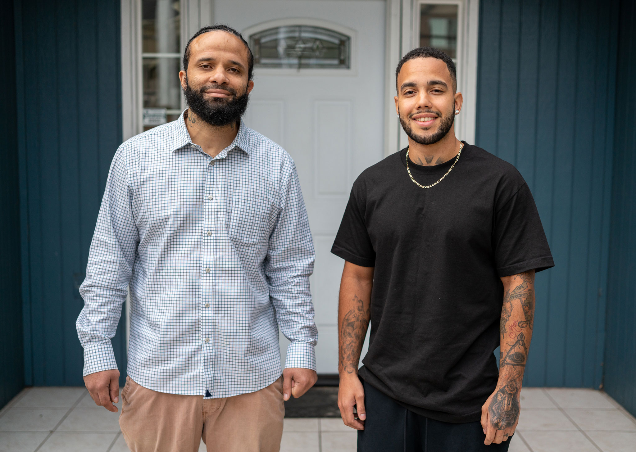 two men stand in front of a teal blue house while smiling proudly.