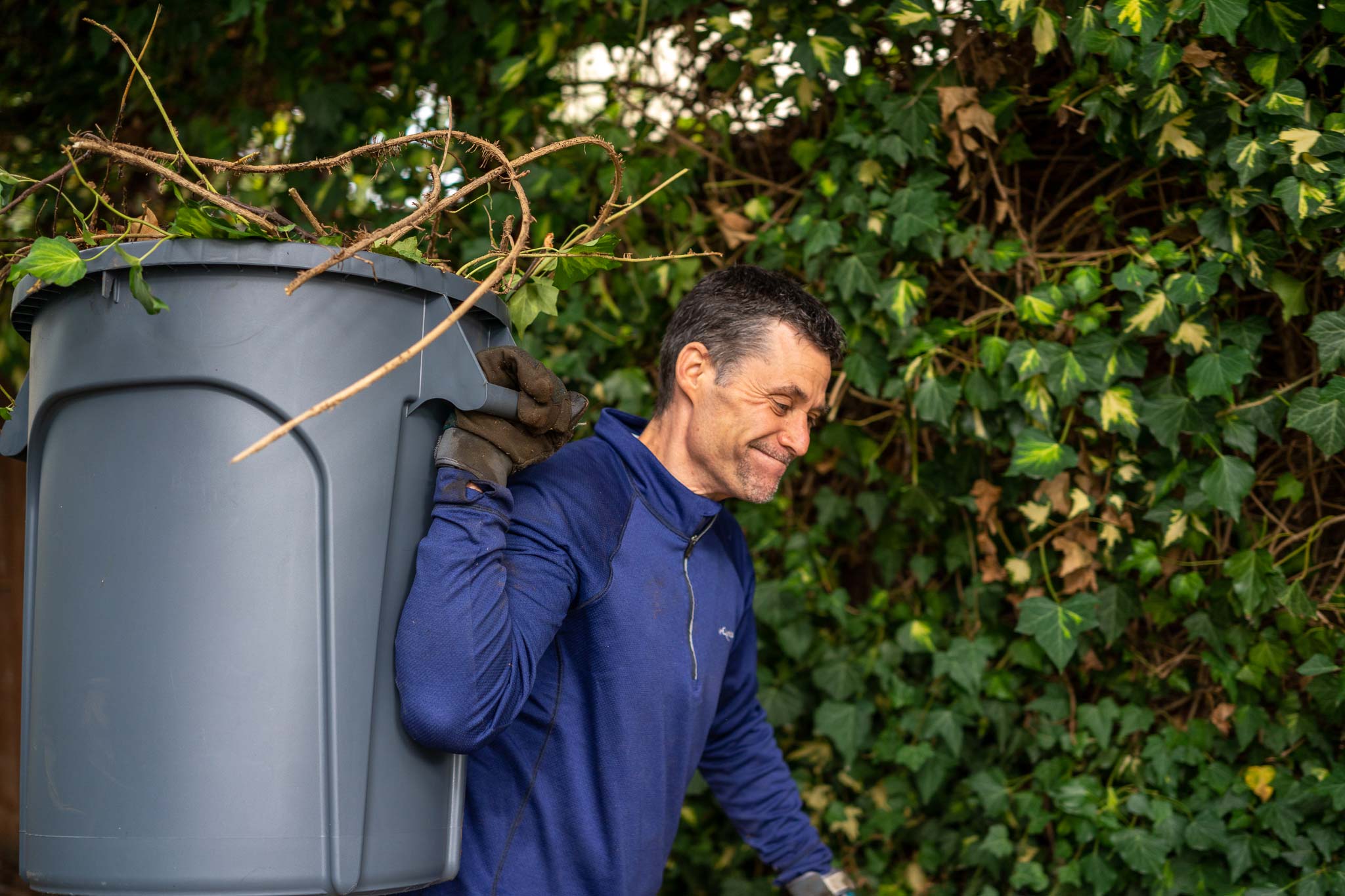 a smiling man wearing gloves holding a full trashcan of vines while moving left to right