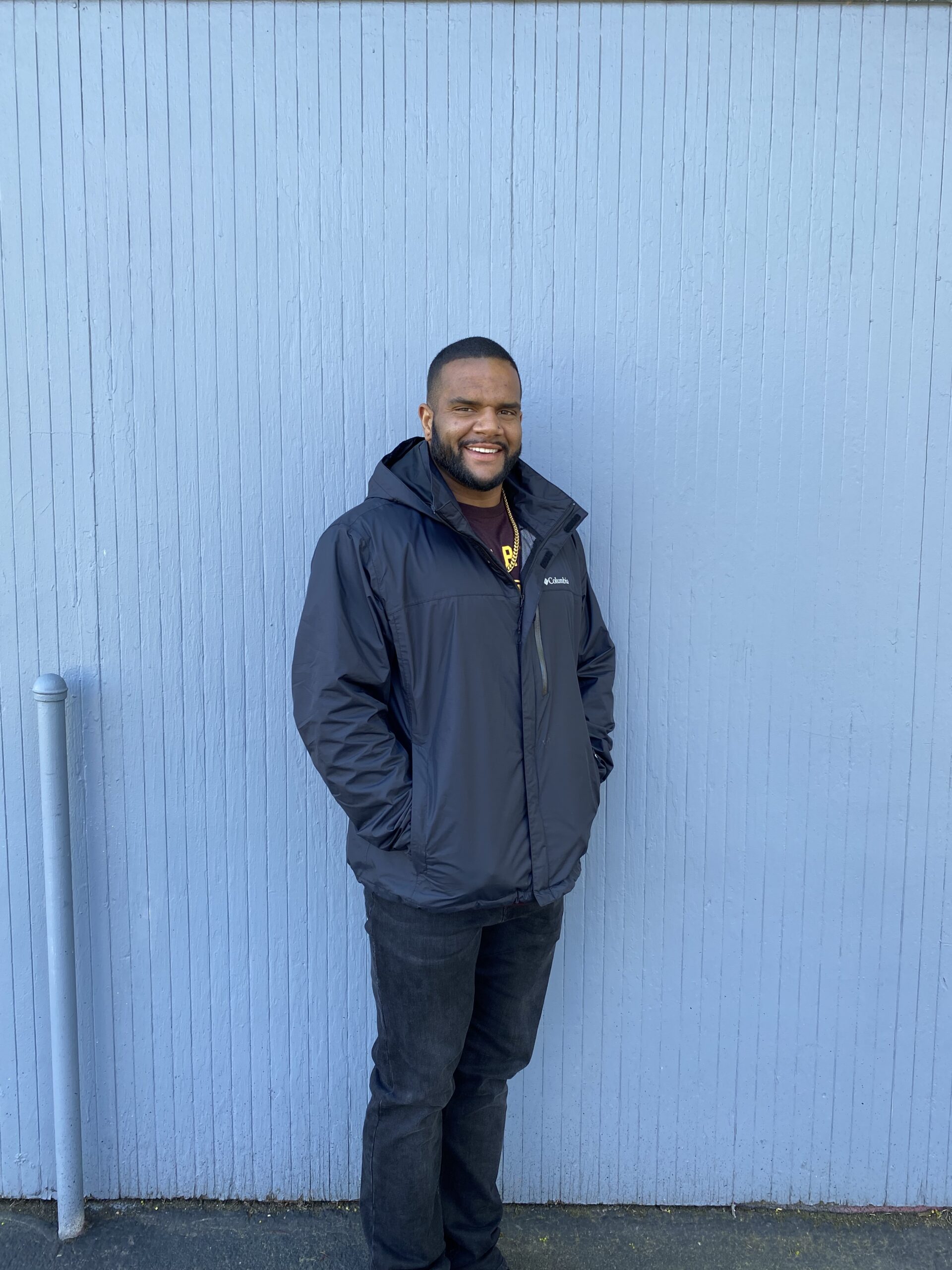 a smiling man with his back against a periwinkle blue wall whose hands are in the pockets of the winter coat he's wearing.