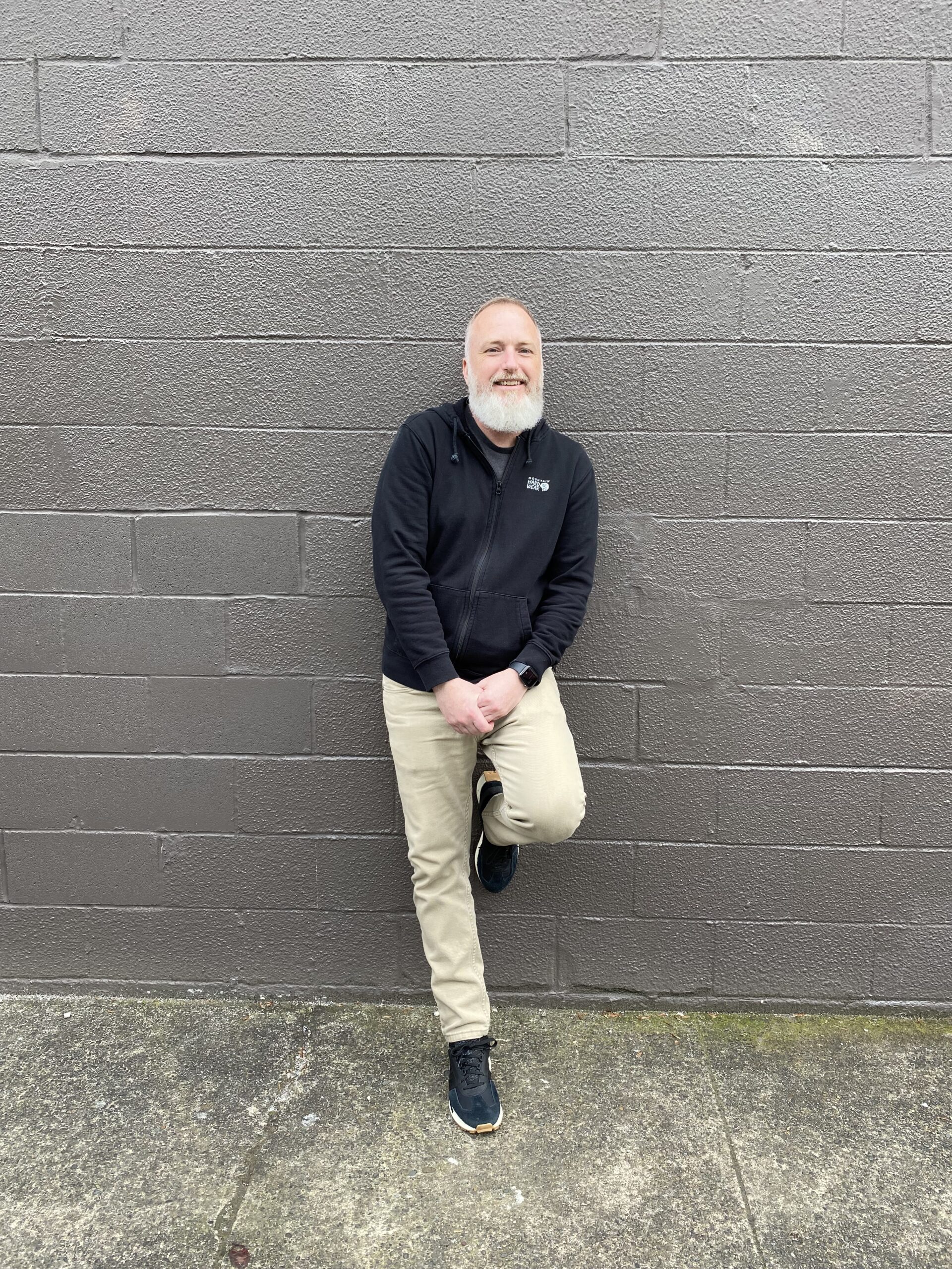 a smiling, gray-bearded man, hands clasped at his waist, standing with one leg up on the black wall behind him