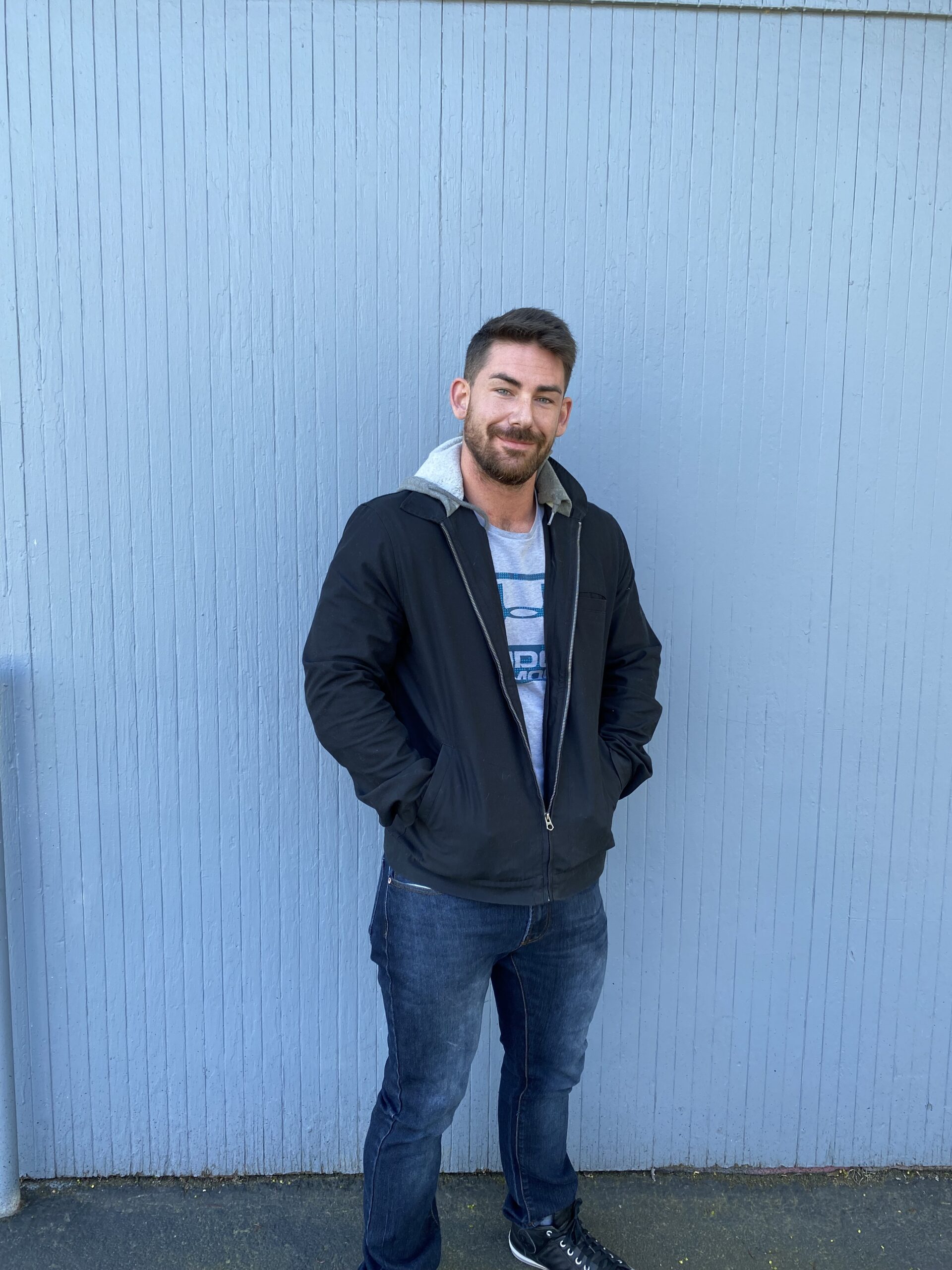 a smiling man with his back against a periwinkle blue wall whose hands are in the pockets of the winter coat he's wearing.