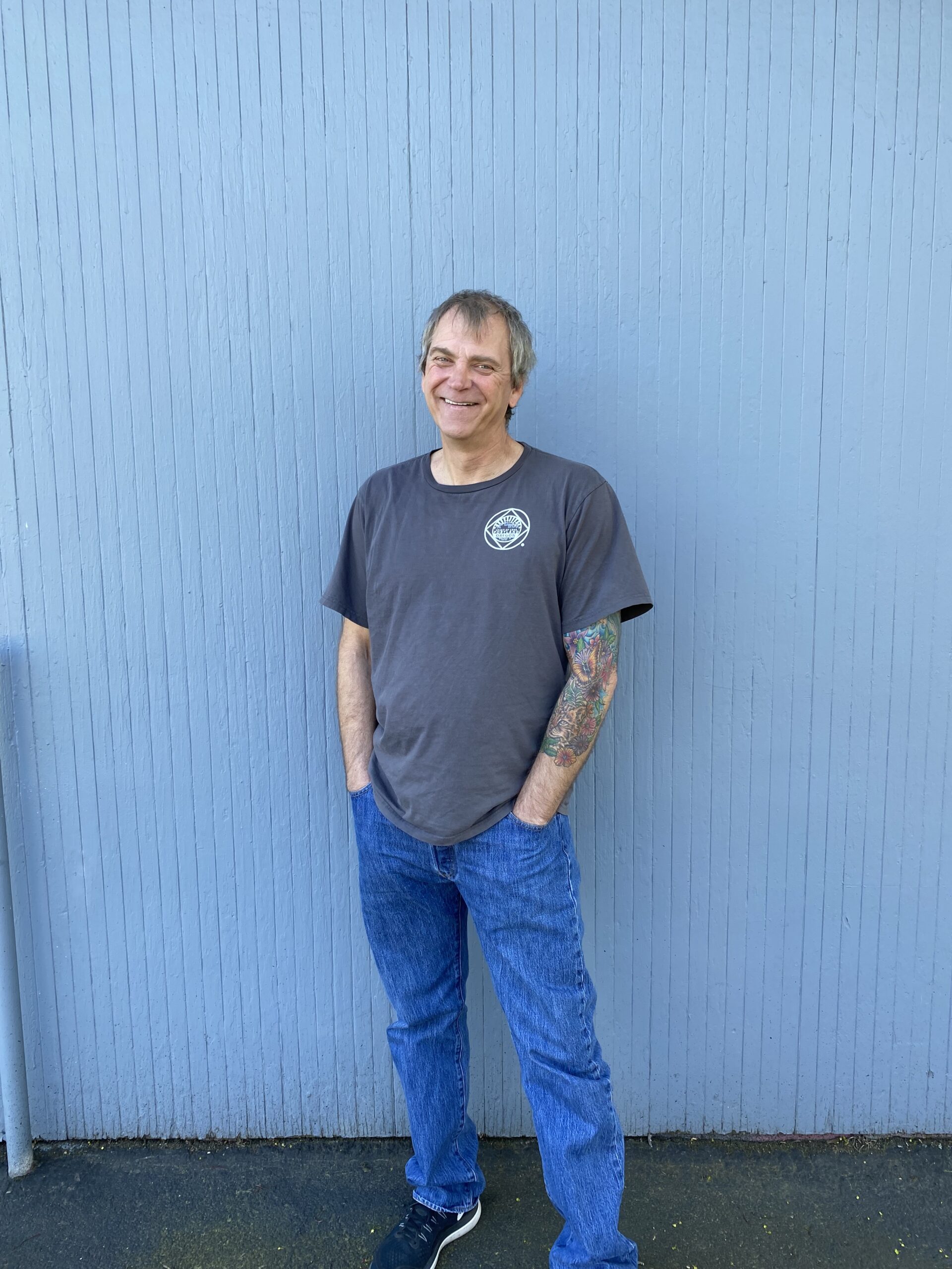 a smiling man, tattoos covering his left arm, with his back against a periwinkle blue wall